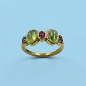 Puede incluir: Un anillo de oro con tres piedras preciosas verdes ovaladas y dos piedras preciosas rojas más pequeñas. El anillo tiene una banda curva y está sobre un fondo azul claro. Las piedras preciosas son el punto focal del anillo.