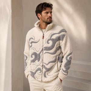Può includere: Una giacca in pile color crema con collo alto e chiusura frontale a zip intera con una piccola linguetta rossa. La giacca è decorata con un intricato motivo grigio a vortice che si estende sul petto e sulle maniche.