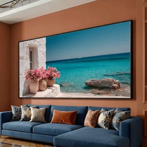 Puede incluir: Gran obra de arte enmarcada que representa una escena costera con agua turquesa y un cielo azul claro. Un edificio blanco con flores rosas en macetas de terracota está a la izquierda. Un sofá seccional azul con cojines decorativos está en primer plano.