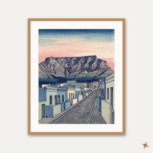 Può includere: Una stampa artistica incorniciata che raffigura una strada acciottolata fiancheggiata da edifici color pastello che conduce verso una grande montagna dalla cima piatta sotto un cielo con sfumature rosa e blu. L'illustrazione mostra una prospettiva.
