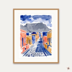 Peut inclure: Une peinture à l'aquarelle encadrée représentant une rue pavée bordée de bâtiments orange, jaunes et bleus. En arrière-plan, une grande montagne grise se dresse sous un ciel bleu foncé. L'œuvre est insérée dans un cadre en bois clair.