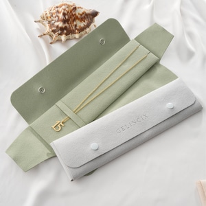 May include: A gold-colored necklace with the number "15" pendant rests inside a light green jewelry case. A light gray jewelry pouch is next to the case. A seashell is in the background. The jewelry case is open, revealing the necklace.
