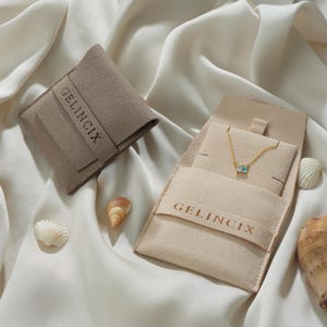 May include: A gold chain necklace with a small rectangular blue and clear stone pendant displayed on a light beige velvet pouch with the text GELINCIX embossed on the front, accompanied by a matching closed pouch and three small seashells.