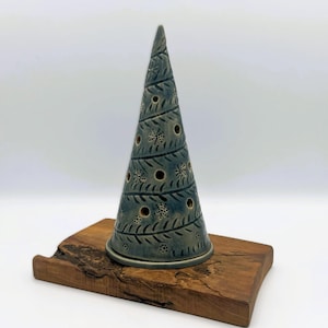 Könnte beinhalten: Eine kegelförmige Keramikskulptur in blaugrauer Glasur mit einem Spiralmuster aus eingravierten farnartigen Blättern und kleinen kreisförmigen Aussparungen. Das Objekt steht auf einem rechteckigen Stück Naturholz mit rauer Kante.