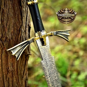 May include: A sword with a patterned Damascus steel blade, a black handle, and a gold-coloured metal crossguard featuring intricate textured details. The logo for VIKINGFINDSSTORE is visible in the upper right corner of the image.