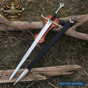 May include: A silver-toned metal sword with a black wrapped handle and a crossguard, resting on a brown wooden wall mount with gold floral patterns. A long, black scabbard with a silver metal tip and throat is positioned diagonally beside the sword.