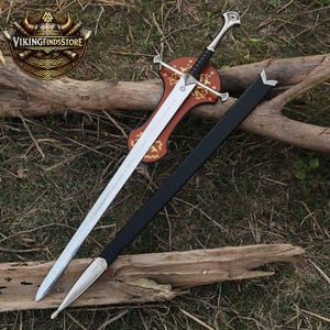 May include: A silver-toned metal sword with a black wrapped handle and a crossguard, featuring an engraved star symbol on the blade. The sword rests on a brown wooden plaque with gold floral patterns, accompanied by a black scabbard with silver accents.