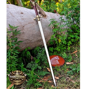 May include: A long sword with a metallic blade, a dark brown cylindrical grip, and an ornate brass crossguard. The sword rests against a large log. Nearby, a circular brown shield features a white graphic of a wolf head in profile.