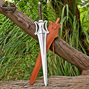 May include: A silver-coloured metal sword with a pointed blade, a complex guard, and a brown wrapped handle. The sword is accompanied by a tan leather sheath featuring visible stitching, metal rivets, and thin leather straps for attachment.