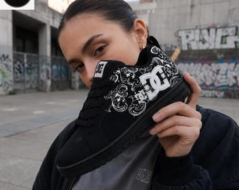 May include: A black trainer featuring a white DC logo on the side and a white intricate swirling pattern across the upper. The shoe has black laces, a padded collar, and a black sole. The tongue displays a black and white DC brand patch.