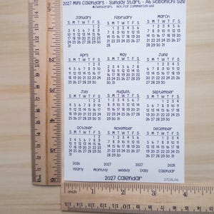 Pu&ograve; includere: Un foglio bianco di minicalendari 2027 con inizio domenica, che misura circa 15,2 cm di altezza e 10,8 cm di larghezza come mostrato dai righelli in legno. Il foglio mostra dodici mesi da gennaio a dicembre con testo nero.