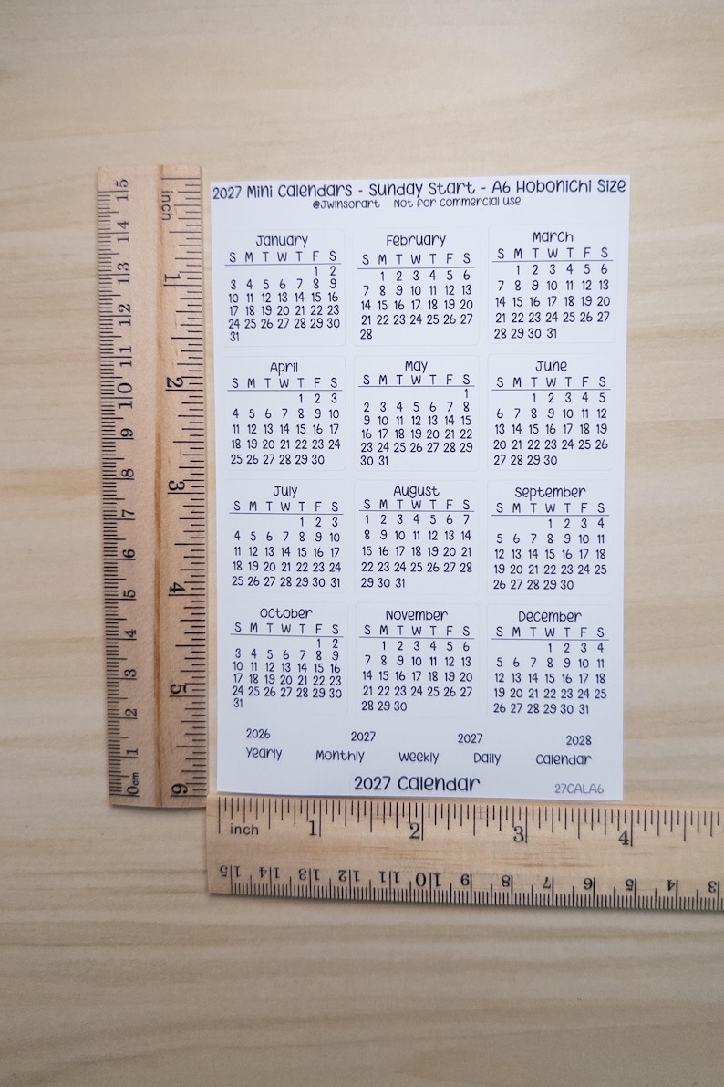 Pu&ograve; includere: Un foglio bianco di minicalendari 2027 con inizio domenica, che misura circa 15,2 cm di altezza e 10,8 cm di larghezza come mostrato dai righelli in legno. Il foglio mostra dodici mesi da gennaio a dicembre con testo nero.