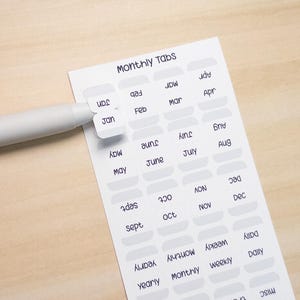 Peut inclure: Feuille d'onglets mensuels blancs avec texte bleu foncé. Les onglets sont étiquetés avec les mois, les jours et d'autres catégories. Un stylo blanc est positionné près de l'onglet "Jan". Le texte "Monthly Tabs" est en haut.