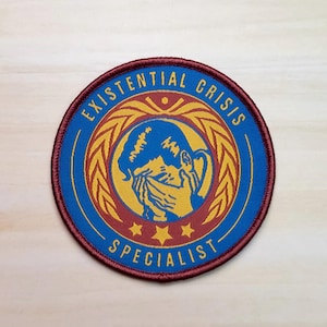 May include: A round, maroon embroidered patch with a blue circle in the center. The blue circle has a gold wreath with a stylized image of a person crying. The text "Existential Crisis Specialist" is written around the outside of the patch.