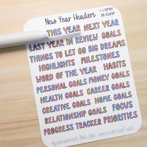 May include: A white sticker sheet with colorful text, including "New Year Headers," "This Year Next Year," and goal-related phrases. A gray pen rests on the sheet. The text is in various colors.