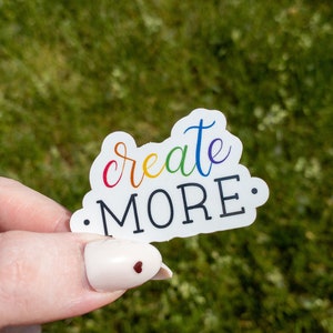 May include: A white sticker with the text "create more." The text is in rainbow colors with black outlines. The sticker is being held by a hand with a white nail with a small red heart design.
