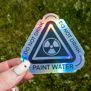 May include: A holographic sticker with a black triangle warning sign. The sign has a radiation symbol inside and the text "DO NOT DRINK PAINT WATER" around the outside.