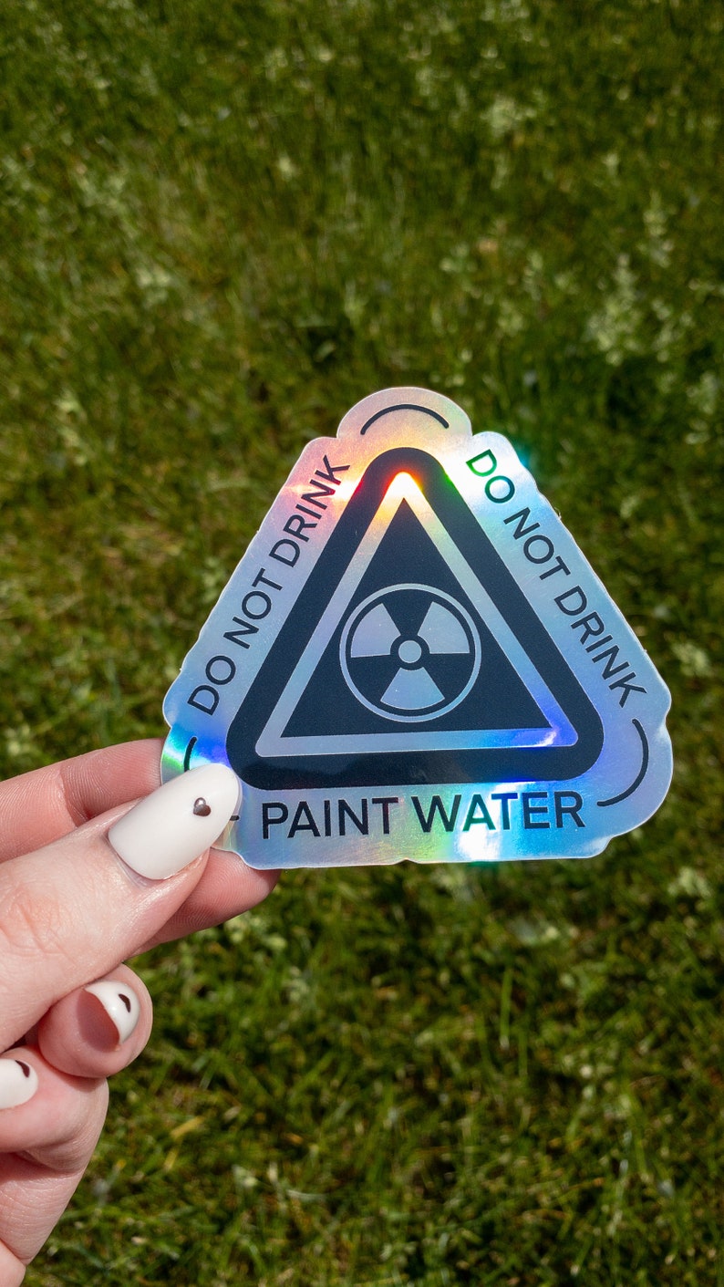 May include: A holographic sticker with a black triangle warning sign. The sign has a radiation symbol inside and the text "DO NOT DRINK PAINT WATER" around the outside.