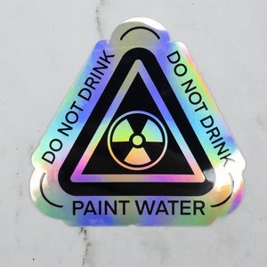 May include: A holographic sticker with a black triangle warning sign. The triangle has a radiation symbol inside and the text "DO NOT DRINK PAINT WATER" around the outside.