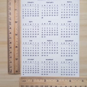 Pu&ograve; includere: Un foglio bianco di minicalendari 2027 con inizio domenica, formato A5 Hobonichi. Il foglio presenta dodici griglie mensili da gennaio a dicembre. Due righelli in legno sono posizionati lungo i bordi sinistro e inferiore per la scala.