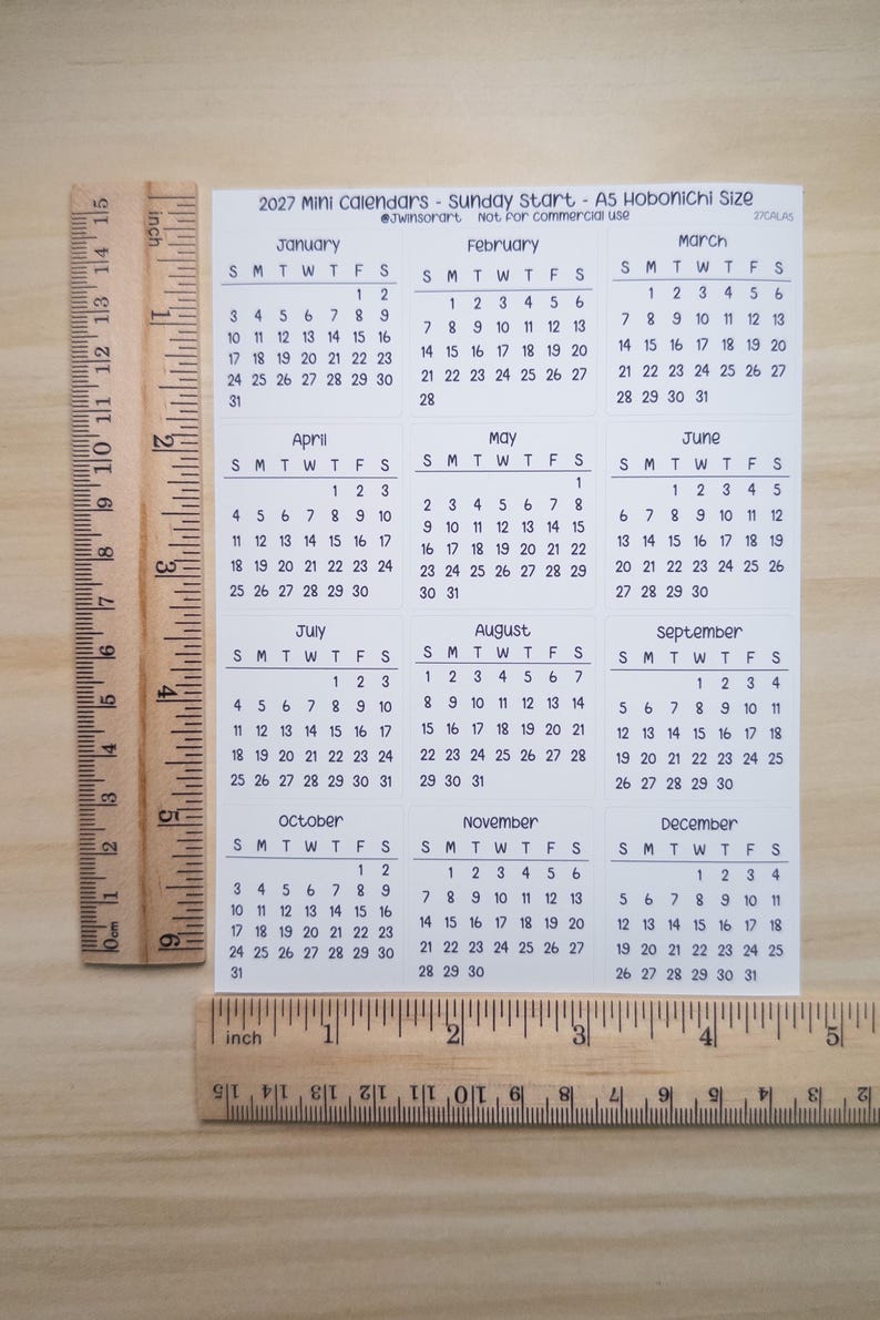 Pu&ograve; includere: Un foglio bianco di minicalendari 2027 con inizio domenica, formato A5 Hobonichi. Il foglio presenta dodici griglie mensili da gennaio a dicembre. Due righelli in legno sono posizionati lungo i bordi sinistro e inferiore per la scala.