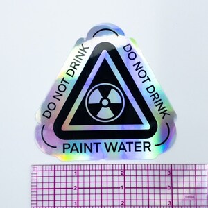 May include: A holographic sticker with a black triangle warning sign. The sign has a radiation symbol inside and the text "DO NOT DRINK PAINT WATER" around the outside.