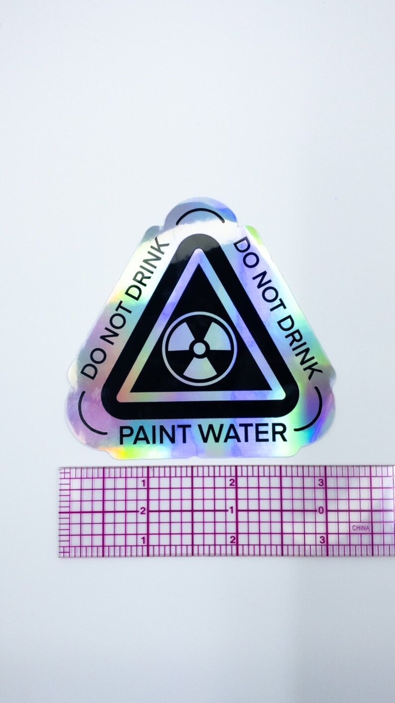 May include: A holographic sticker with a black triangle warning sign. The sign has a radiation symbol inside and the text "DO NOT DRINK PAINT WATER" around the outside.