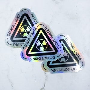 May include: Three iridescent holographic stickers with a black triangle warning sign. The sign has a black radiation symbol and the text "DO NOT DRINK PAINT WATER" around the triangle.