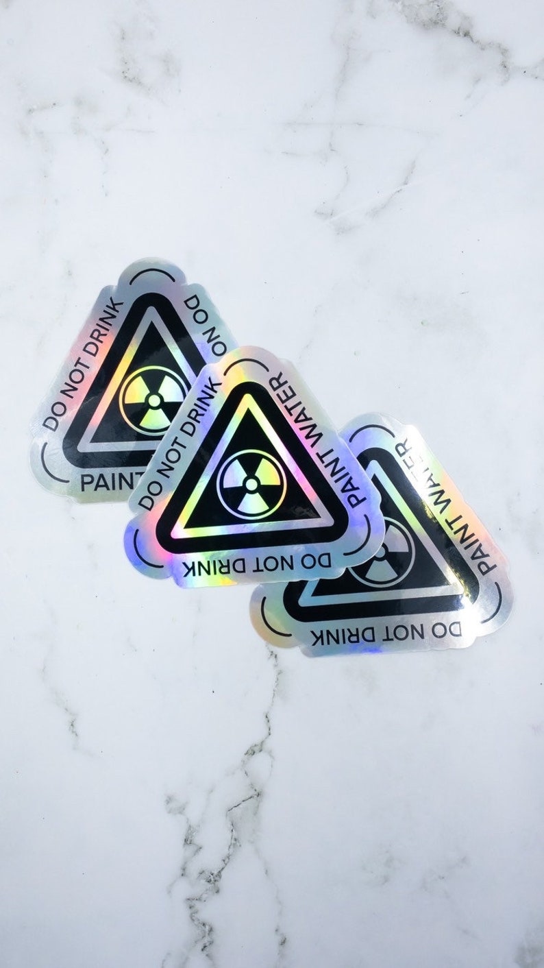 May include: Three iridescent holographic stickers with a black triangle warning sign. The sign has a black radiation symbol and the text "DO NOT DRINK PAINT WATER" around the triangle.