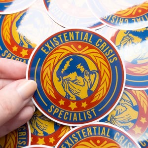 May include: A close-up of several round stickers with a blue, yellow, and red design. The stickers feature a cartoon illustration of a person crying with the text "Existential Crisis Specialist" in a yellow banner.