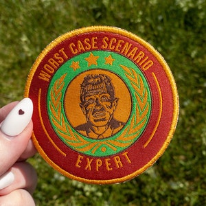 May include: A red, yellow, and green embroidered patch with a cartoon man's face and the text "Worst Case Scenario Expert".