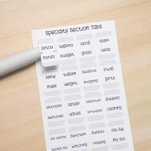 May include: A sheet of specialty section tabs with various labels like "notes," "budget," and "goals." The tabs are white with gray borders and black text. A white pen points to the "notes" tab. The background is a light wood surface.