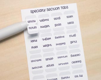 Könnte beinhalten: Ein Bogen mit Spezialabschnitts-Tabs mit Etiketten wie "Notizen", "Budget" und "Ziele". Die Tabs sind weiß mit grauen Rändern und schwarzem Text. Ein weißer Stift zeigt auf den Tab "Notizen". Der Hintergrund ist eine helle Holzoberfläche.