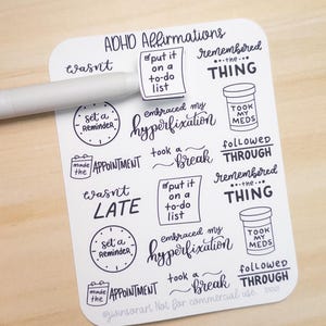 May include: A white paper with handwritten "ADHD Affirmations" in black ink. Affirmations include "Set a Reminder," "Took my meds," and "Put it on a to-do list." A white pen rests on the paper.
