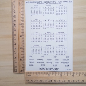 Pu&ograve; includere: Un foglio rettangolare bianco con un mini calendario 2027 che inizia di domenica, posizionato accanto a righelli in legno con misure in pollici e centimetri. Il foglio include griglie mensili da gennaio a dicembre ed etichette di testo.