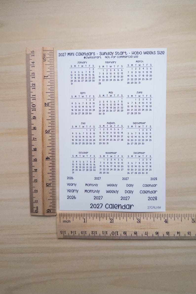 Pu&ograve; includere: Un foglio rettangolare bianco con un mini calendario 2027 che inizia di domenica, posizionato accanto a righelli in legno con misure in pollici e centimetri. Il foglio include griglie mensili da gennaio a dicembre ed etichette di testo.