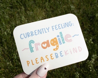 May include: A white rectangular card with rounded corners, held against a green grassy background. The card features the text "CURRENTLY FEELING: FRAGILE PLEASE BE KIND" in various colorful fonts. The word "fragile" has a heart icon.