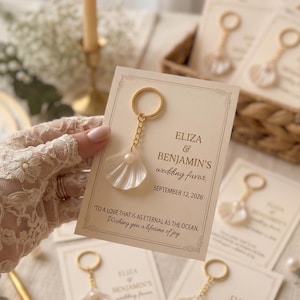 Puede incluir: Un llavero de metal dorado con un dije de concha blanca y una pequeña perla, sujeto a una tarjeta rectangular de color crema. La tarjeta muestra el texto: ELIZA & BENJAMIN'S wedding favor, SEPTEMBER 12, 2026, TO A LOVE THAT IS AS ETERNAL AS THE OCEAN.