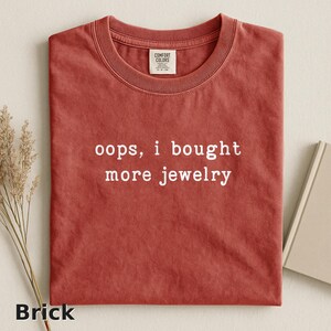 May include: Brick-colored Comfort Colors t-shirt with the phrase "oops, i bought more jewelry" in white lettering. The shirt has a crew neck and short sleeves. A small tag is visible near the collar.