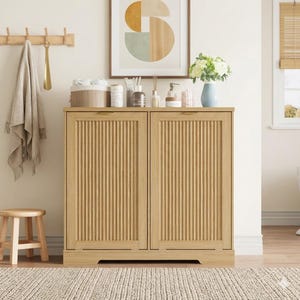 May include: A light-colored wooden cabinet with two doors featuring vertical grooved detailing. The cabinet is part of a bathroom or bedroom set, with a small stool and a rug in front. A framed artwork and decorative items sit on top.