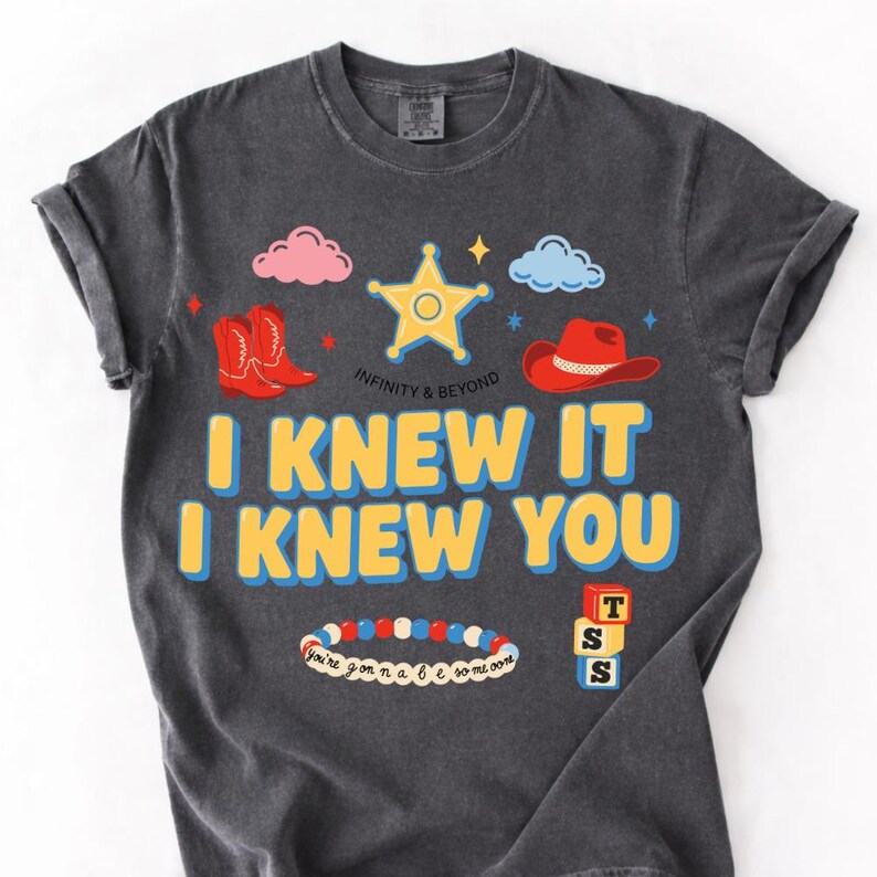 May include: A charcoal gray short-sleeve t-shirt featuring a graphic print with a yellow sheriff star, red cowboy boots, a red cowboy hat, clouds, a beaded bracelet, and stacked blocks. The text reads I KNEW IT I KNEW YOU and INFINITY & BEYOND.