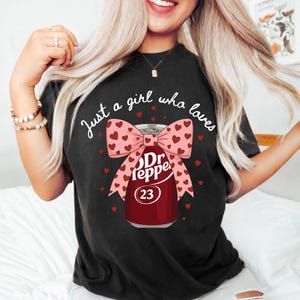 Peut inclure: Un t-shirt noir avec un motif représentant une canette de soda bordeaux ornée d'un grand nœud rose à petits cœurs rouges. Le texte "Just a girl who loves" est imprimé en blanc au-dessus de la canette, affichant le logo Dr Pepper et le chiffre 23.