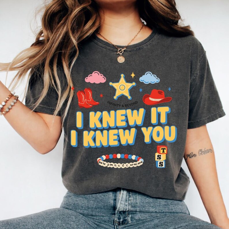 May include: A charcoal gray t-shirt featuring a graphic print with a yellow sheriff star, red cowboy boots, a red cowboy hat, pink and blue clouds, a beaded bracelet, and stacked alphabet blocks. The text reads I KNEW IT I KNEW YOU.