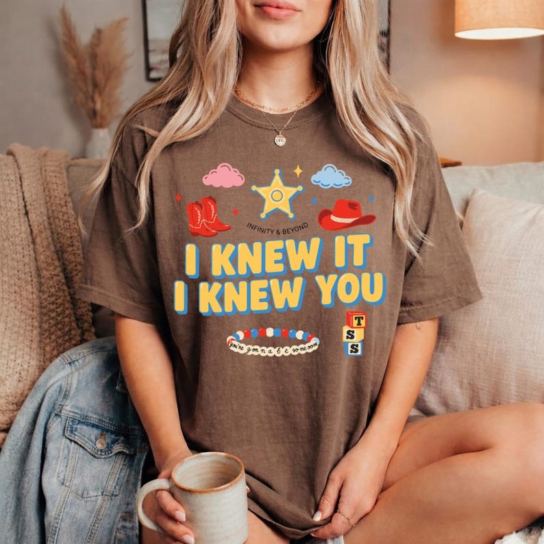 May include: A brown short-sleeve t-shirt featuring a graphic print with a yellow sheriff star, red cowboy boots, a red cowboy hat, pink and blue clouds, a colorful beaded bracelet, and three stacked blocks with the letters T, S, and S. The text reads I KNEW IT I KNEW YOU.