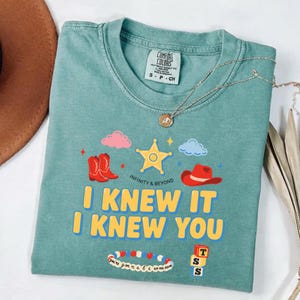 May include: A sage green crewneck t-shirt featuring a graphic print with a yellow sheriff star, red cowboy boots, a red cowboy hat, pink and blue clouds, a beaded bracelet, and three stacked blocks with the letters T, S, S. Text reads I KNEW IT I KNEW YOU.