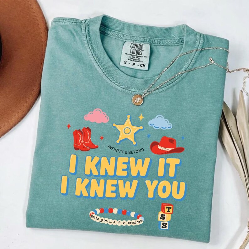 May include: A sage green crewneck t-shirt featuring a graphic print with a yellow sheriff star, red cowboy boots, a red cowboy hat, pink and blue clouds, a beaded bracelet, and three stacked blocks with the letters T, S, S. Text reads I KNEW IT I KNEW YOU.