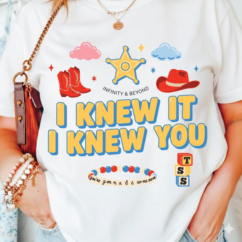 May include: A white t-shirt featuring a graphic print with a yellow sheriff star, red cowboy boots, a red cowboy hat, and blue and pink clouds. The text reads "INFINITY & BEYOND", "I KNEW IT I KNEW YOU", and "You're gonna be someone" with toy blocks.
