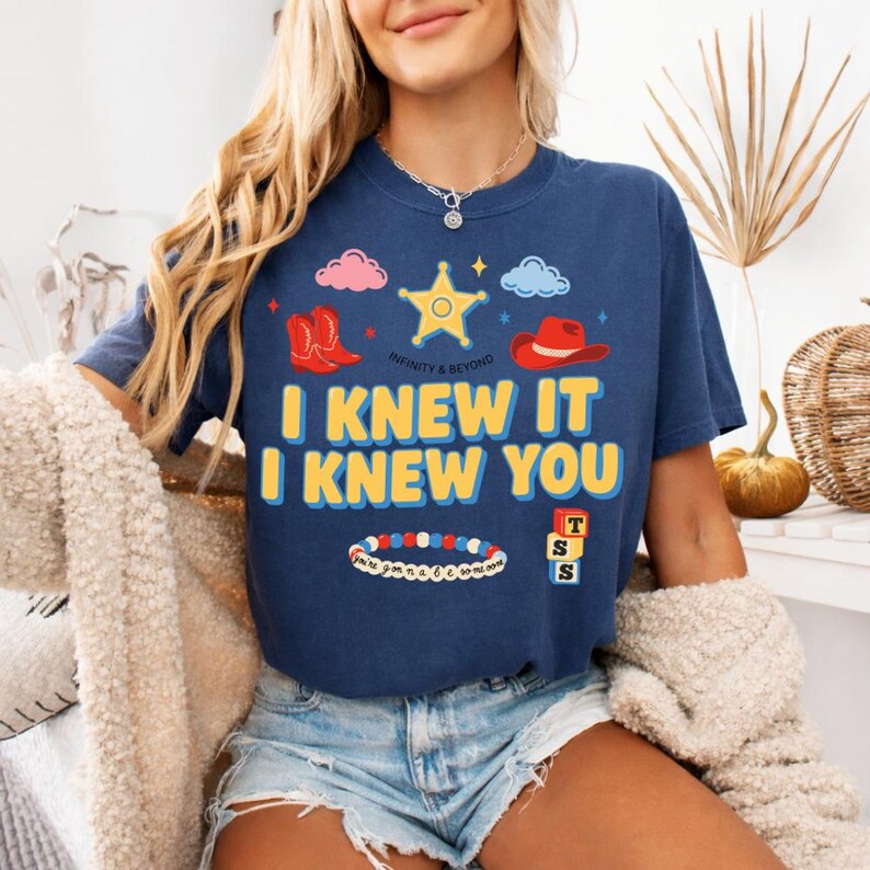 May include: A navy blue short-sleeve t-shirt featuring a graphic print with a yellow sheriff star, red cowboy boots, a red cowboy hat, a beaded bracelet, and alphabet blocks. The text reads I KNEW IT I KNEW YOU and INFINITY & BEYOND.