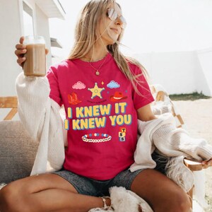 May include: A bright pink short-sleeved t-shirt featuring a graphic print with a yellow sheriff star, red cowboy boots, a red hat, a beaded bracelet, and stacked blocks. The text reads I KNEW IT I KNEW YOU in bold yellow letters with blue outlines.