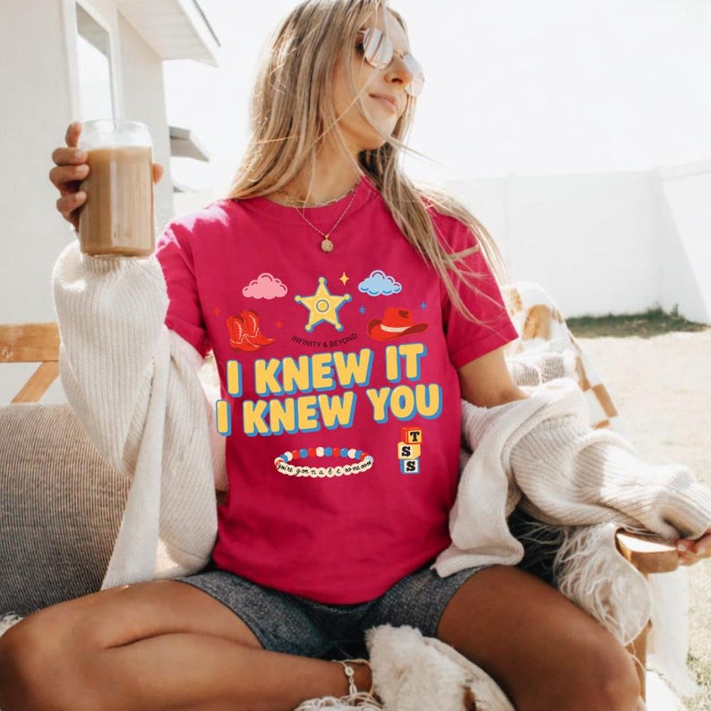 May include: A bright pink short-sleeved t-shirt featuring a graphic print with a yellow sheriff star, red cowboy boots, a red hat, a beaded bracelet, and stacked blocks. The text reads I KNEW IT I KNEW YOU in bold yellow letters with blue outlines.